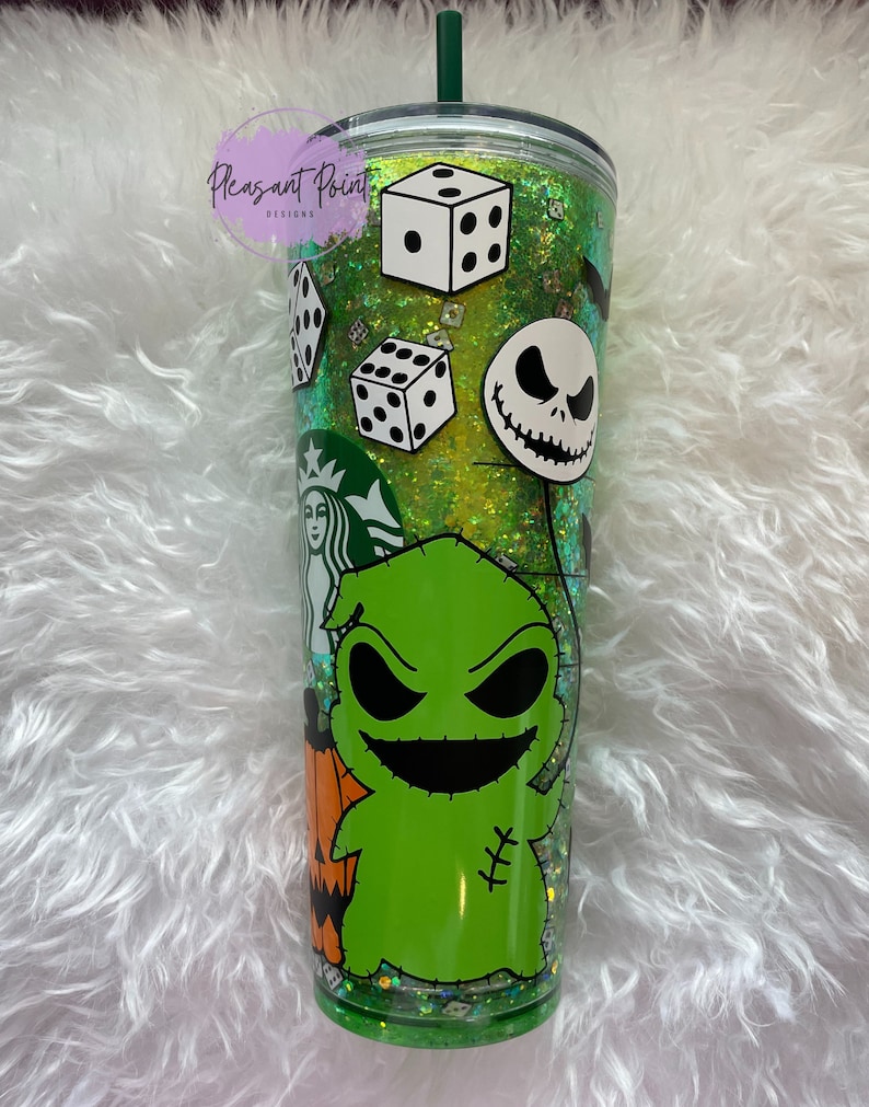 Puede incluir: Un vaso transparente de doble pared con un interior de purpurina verde, con calcoman&iacute;as de personajes de Pesadilla antes de Navidad. Incluye im&aacute;genes de dados, Jack Skellington y Oogie Boogie. El vaso tiene una pajita verde y el texto "Pleasant Point Designs".