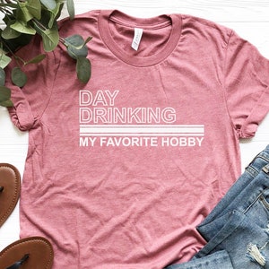 K&ouml;nnte beinhalten: Ein pinkfarbenes T-Shirt mit dem Text "DAY DRINKING MY FAVORITE HOBBY" in Wei&szlig;. Das Shirt ist gefaltet und liegt auf einer wei&szlig;en Oberfl&auml;che mit einem Paar braunen Sandalen und einer blauen Jeans.