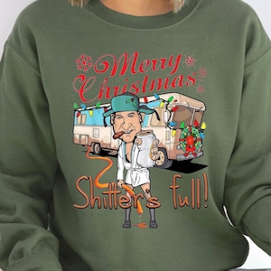 May include: Green Christmas sweatshirt with a cartoon illustration of a man in a white robe holding a mug, standing in front of a camper van decorated with Christmas lights and a wreath. The text "Merry Christmas" and "Shitter's Full!" is printed on the sweatshirt.