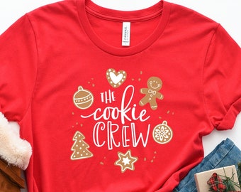 May include: Red t-shirt with the text "The cookie crew" in white lettering. The shirt features a gingerbread man, a heart, a Christmas tree, and ornaments.