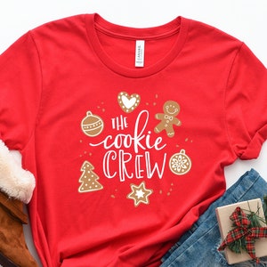 May include: Red t-shirt with the text "The cookie crew" in white lettering. The shirt features a gingerbread man, a heart, a Christmas tree, and ornaments.