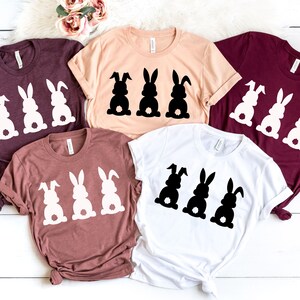 Pode incluir: Quatro camisetas de diferentes cores e designs com tr&ecirc;s silhuetas de coelhos. Os coelhos est&atilde;o todos olhando na mesma dire&ccedil;&atilde;o e t&ecirc;m uma forma de cora&ccedil;&atilde;o na parte de tr&aacute;s. As camisetas est&atilde;o todas dobradas e dispostas sobre uma superf&iacute;cie de madeira branca.