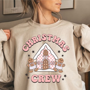 May include: A beige sweatshirt with a pink graphic design of a gingerbread house, two gingerbread people, and the text "Christmas Crew".