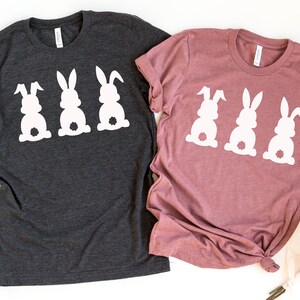 Pode incluir: Duas camisetas com um design de tr&ecirc;s silhuetas de coelhos brancos. A camiseta &agrave; esquerda &eacute; cinza escuro e a camiseta &agrave; direita &eacute; rosa poeira.