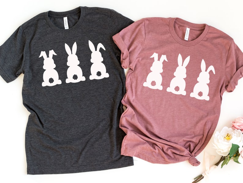 Pode incluir: Duas camisetas com um design de tr&ecirc;s silhuetas de coelhos brancos. A camiseta &agrave; esquerda &eacute; cinza escuro e a camiseta &agrave; direita &eacute; rosa poeira.