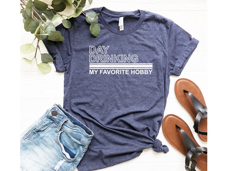 K&ouml;nnte beinhalten: Ein marineblaues T-Shirt mit wei&szlig;em Text, der "DAY DRINKING MY FAVORITE HOBBY" lautet.