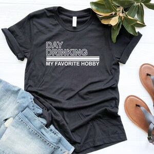 K&ouml;nnte beinhalten: Schwarzes T-Shirt mit wei&szlig;em Text, der "Day Drinking My Favorite Hobby" lautet.