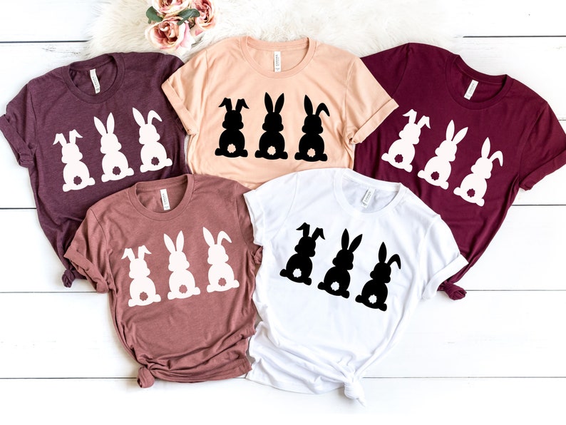 Pode incluir: Quatro camisetas de diferentes cores e designs com tr&ecirc;s silhuetas de coelhos. Os coelhos est&atilde;o todos olhando na mesma dire&ccedil;&atilde;o e t&ecirc;m uma forma de cora&ccedil;&atilde;o na parte de tr&aacute;s. As camisetas est&atilde;o todas dobradas e dispostas sobre uma superf&iacute;cie de madeira branca.