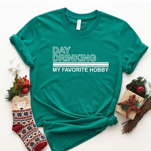 K&ouml;nnte beinhalten: Ein gr&uuml;nes T-Shirt mit wei&szlig;em Text, der "DAY DRINKING MY FAVORITE HOBBY" lautet.