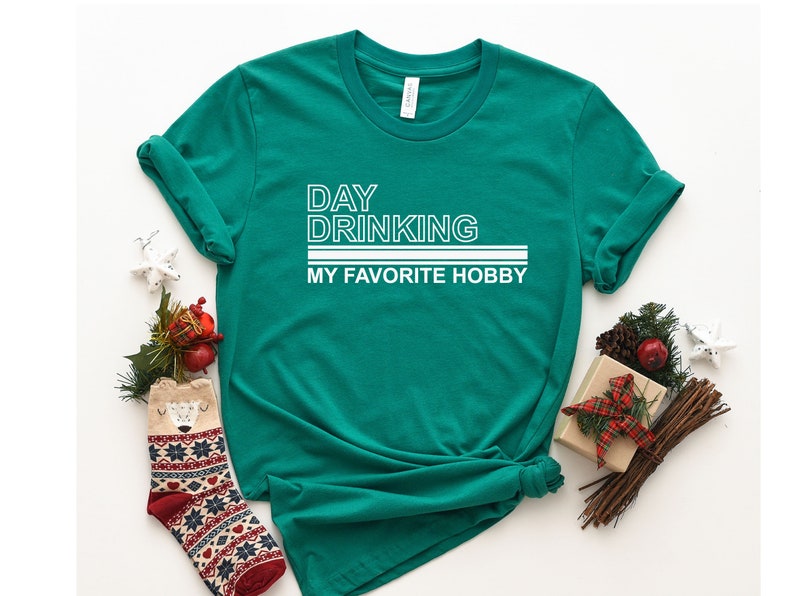 K&ouml;nnte beinhalten: Ein gr&uuml;nes T-Shirt mit wei&szlig;em Text, der "DAY DRINKING MY FAVORITE HOBBY" lautet.