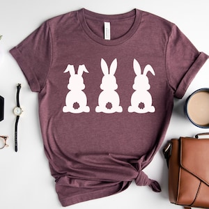Pode incluir: Uma camiseta marrom com um gr&aacute;fico branco de tr&ecirc;s silhuetas de coelhos. Os coelhos est&atilde;o de costas para o espectador com suas caudas vis&iacute;veis.
