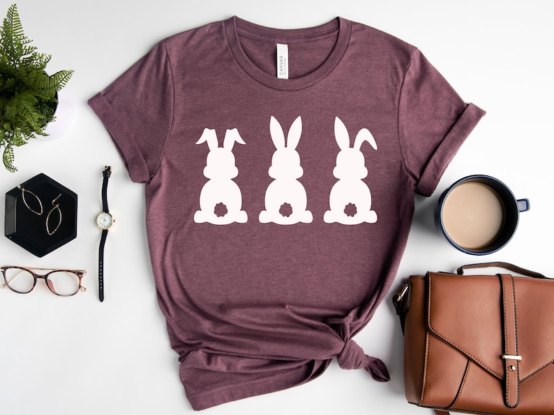 Pode incluir: Uma camiseta marrom com um gr&aacute;fico branco de tr&ecirc;s silhuetas de coelhos. Os coelhos est&atilde;o de costas para o espectador com suas caudas vis&iacute;veis.