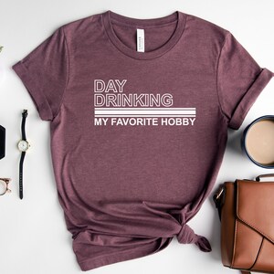 K&ouml;nnte beinhalten: Ein maronfarbenes T-Shirt mit wei&szlig;em Text, der "Day Drinking My Favorite Hobby" lautet.