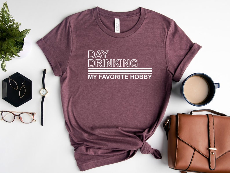 K&ouml;nnte beinhalten: Ein maronfarbenes T-Shirt mit wei&szlig;em Text, der "Day Drinking My Favorite Hobby" lautet.