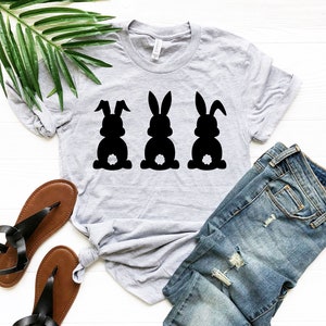 Pode incluir: Uma camiseta cinza com um gr&aacute;fico de tr&ecirc;s silhuetas de coelhos pretos. Os coelhos est&atilde;o olhando para a frente com suas caudas levantadas. A camiseta est&aacute; colocada sobre uma superf&iacute;cie branca com um par de sand&aacute;lias marrons e uma cal&ccedil;a jeans azul.