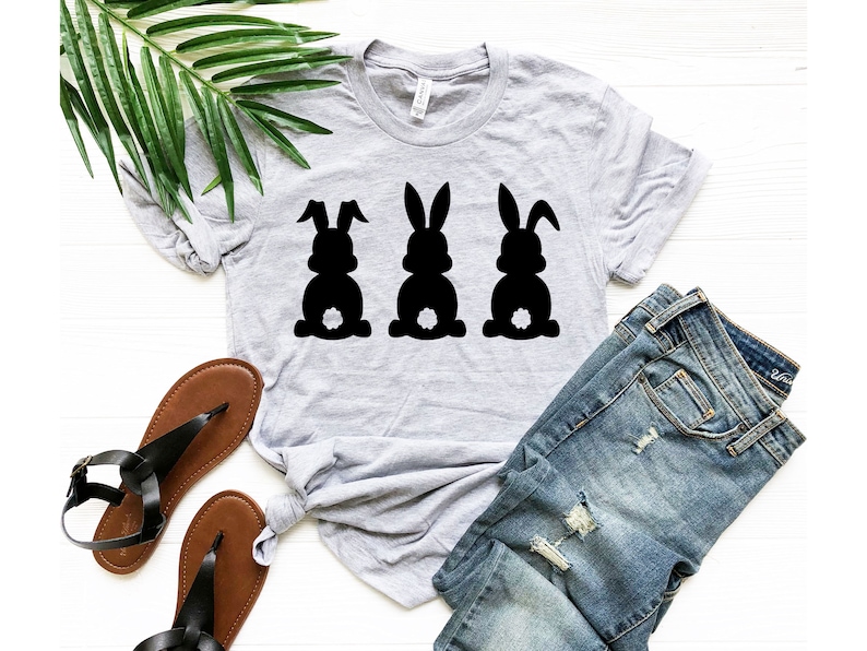 Pode incluir: Uma camiseta cinza com um gr&aacute;fico de tr&ecirc;s silhuetas de coelhos pretos. Os coelhos est&atilde;o olhando para a frente com suas caudas levantadas. A camiseta est&aacute; colocada sobre uma superf&iacute;cie branca com um par de sand&aacute;lias marrons e uma cal&ccedil;a jeans azul.
