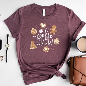 May include: A maroon t-shirt with the text "The cookie crew" in white lettering surrounded by gingerbread cookie-themed illustrations.