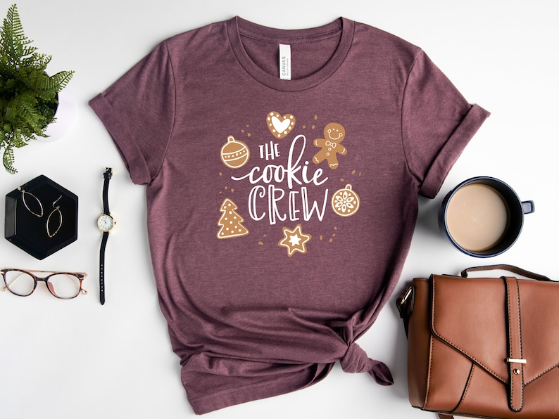 May include: A maroon t-shirt with the text "The cookie crew" in white lettering surrounded by gingerbread cookie-themed illustrations.