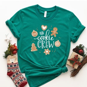 May include: A green t-shirt with the text "The Cookie Crew" in white lettering. The shirt features a gingerbread man, a heart, a Christmas tree, a star, and an ornament.