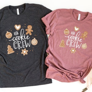 May include: Two t-shirts with the text "The Cookie Crew" in white lettering. The shirts are decorated with gingerbread cookies, ornaments, and stars. One shirt is dark gray and the other is a dusty rose color.