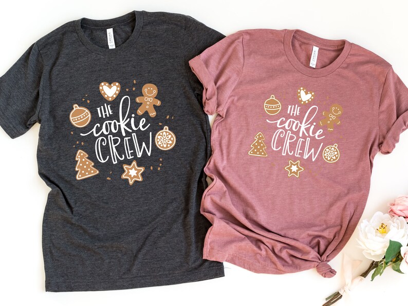 May include: Two t-shirts with the text "The Cookie Crew" in white lettering. The shirts are decorated with gingerbread cookies, ornaments, and stars. One shirt is dark gray and the other is a dusty rose color.