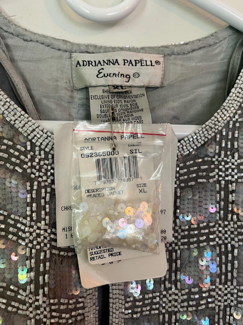 Puede incluir: Chaqueta con cuentas Adrianna Papell Evening plateada, con un patr&oacute;n de cuadr&iacute;cula de lentejuelas y cuentas. La etiqueta dice "Beaded Jacket" y "XL". La chaqueta tiene una capa de seda y forro de ray&oacute;n. Se adjunta una peque&ntilde;a bolsa de lentejuelas.