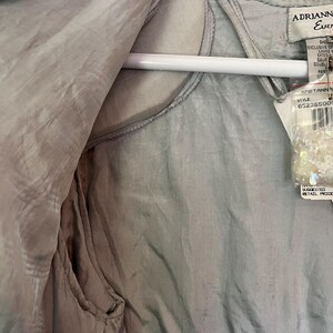Puede incluir: Una prenda de color gris claro y sedosa, posiblemente un chaleco o una blusa, cuelga de una percha blanca. La tela tiene un brillo sutil y parece estar forrada. Una etiqueta con la marca "Adrianna Ever" y un n&uacute;mero de estilo est&aacute; adjunta.