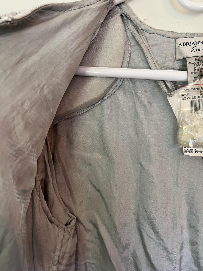 Puede incluir: Una prenda de color gris claro y sedosa, posiblemente un chaleco o una blusa, cuelga de una percha blanca. La tela tiene un brillo sutil y parece estar forrada. Una etiqueta con la marca "Adrianna Ever" y un n&uacute;mero de estilo est&aacute; adjunta.