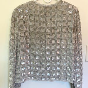Puede incluir: Chaqueta plateada con lentejuelas y un patr&oacute;n de cuadr&iacute;cula. La chaqueta de manga larga est&aacute; cubierta de peque&ntilde;as lentejuelas iridiscentes, creando un efecto brillante. La chaqueta est&aacute; colgada en una percha blanca contra una pared de color claro.