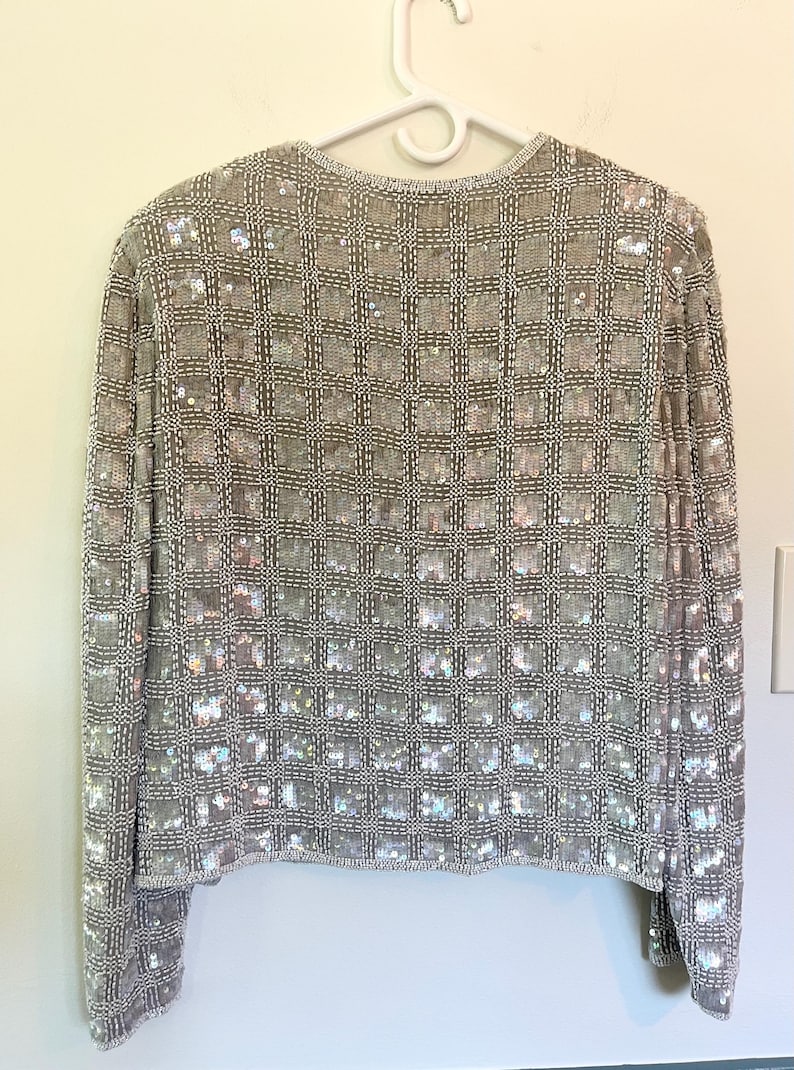 Puede incluir: Chaqueta plateada con lentejuelas y un patr&oacute;n de cuadr&iacute;cula. La chaqueta de manga larga est&aacute; cubierta de peque&ntilde;as lentejuelas iridiscentes, creando un efecto brillante. La chaqueta est&aacute; colgada en una percha blanca contra una pared de color claro.