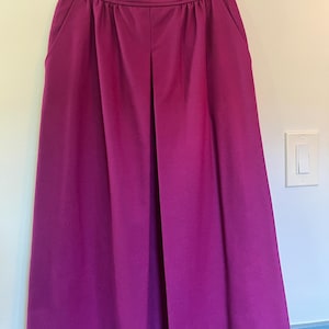May include: A pair of magenta-colored, pleated skirt hanging on a black hanger. The skirt has a high waist, two side pockets, and a button closure. The fabric appears to be a smooth, solid color, and the skirt is knee-length.