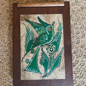 May include: A wooden box with a hinged lid, featuring a hand-painted illustration of a green bird perched on a green floral stem. The bird and floral details are accented with yellow and white on a textured, tan background.