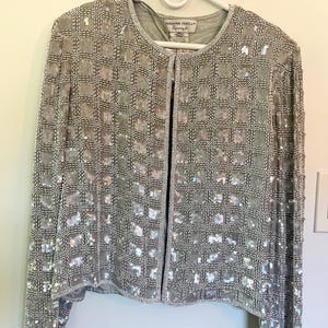 Puede incluir: Chaqueta plateada con cuentas con un patr&oacute;n de cuadr&iacute;cula, colgada de una percha blanca. La chaqueta tiene mangas largas y cuello redondo. La etiqueta dice "Adrianna Papell Evening".