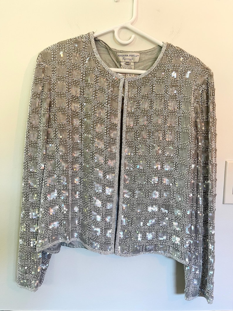 Puede incluir: Chaqueta plateada con cuentas con un patr&oacute;n de cuadr&iacute;cula, colgada de una percha blanca. La chaqueta tiene mangas largas y cuello redondo. La etiqueta dice "Adrianna Papell Evening".