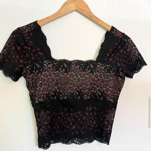 May include: Black lace crop top with a square neckline and short sleeves. The top features a pattern of red shapes on a nude background. The garment is hanging on a wooden hanger against a white wall.