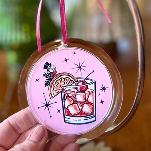 Pu&ograve; includere: Addobbo natalizio rosa con un fronte in vetro e uno sfondo rosa. L'addobbo presenta un'illustrazione di cartone animato di un cocktail con una ciliegia, una fetta d'arancia e cubetti di ghiaccio. Ci sono anche stelle e un pupazzo di neve sull'addobbo.