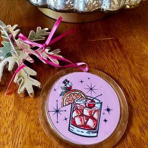 Pu&ograve; includere: Addobbo natalizio rotondo rosa con una copertura in plastica trasparente. L'addobbo presenta un'illustrazione retr&ograve; di un cocktail con una ciliegia, una fetta d'arancia e cubetti di ghiaccio. Uno piccolo pupazzo di neve si trova sullo sfondo.