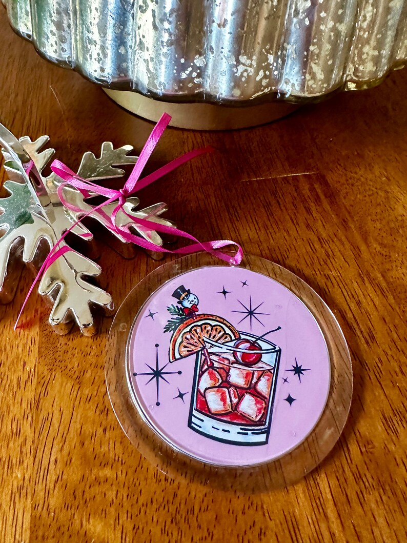 Pu&ograve; includere: Addobbo natalizio rotondo rosa con una copertura in plastica trasparente. L'addobbo presenta un'illustrazione retr&ograve; di un cocktail con una ciliegia, una fetta d'arancia e cubetti di ghiaccio. Uno piccolo pupazzo di neve si trova sullo sfondo.