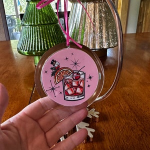 Pu&ograve; includere: Addobbo natalizio rotondo rosa con una copertura in plastica trasparente. L'addobbo presenta un'illustrazione retr&ograve; di un cocktail con una ciliegia, una fetta d'arancia e cubetti di ghiaccio. Lo sfondo &egrave; rosa con stelle bianche.