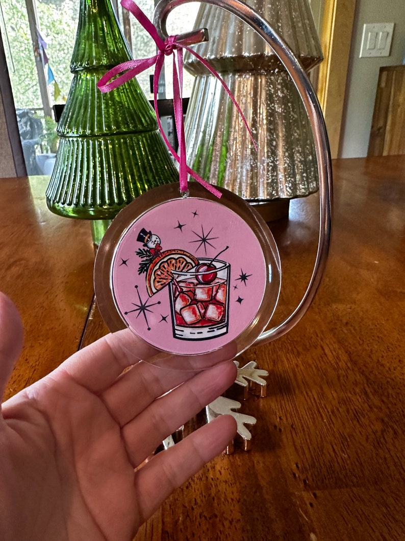 Pu&ograve; includere: Addobbo natalizio rotondo rosa con una copertura in plastica trasparente. L'addobbo presenta un'illustrazione retr&ograve; di un cocktail con una ciliegia, una fetta d'arancia e cubetti di ghiaccio. Lo sfondo &egrave; rosa con stelle bianche.