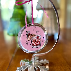 Pu&ograve; includere: Un ornamento natalizio rosa e argento con un'illustrazione vintage di un cocktail con una ciliegia e una fetta d'arancia. L'ornamento &egrave; appeso a un supporto a forma di fiocco di neve argentato.