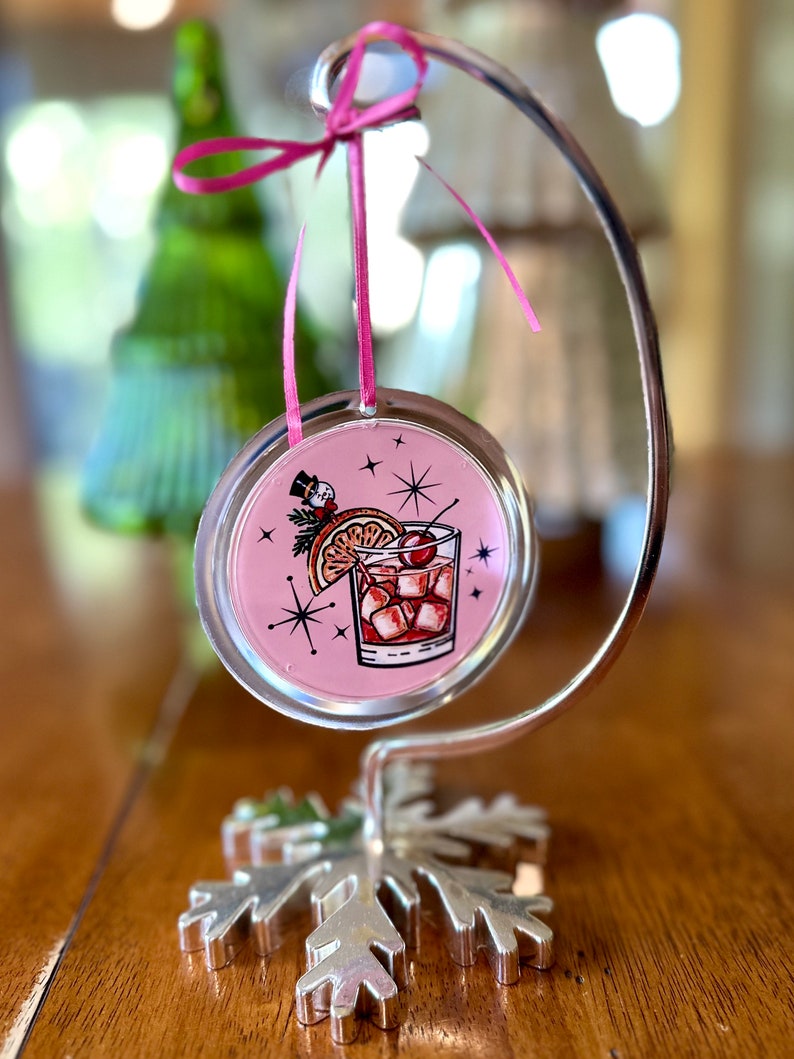 Pu&ograve; includere: Un ornamento natalizio rosa e argento con un'illustrazione vintage di un cocktail con una ciliegia e una fetta d'arancia. L'ornamento &egrave; appeso a un supporto a forma di fiocco di neve argentato.