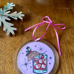 Pu&ograve; includere: Un addobbo natalizio rotondo rosa e trasparente con un'illustrazione di cocktail festivo. Il cocktail &egrave; una bevanda rossa con cubetti di ghiaccio, una ciliegia, una fetta d'arancia e un rametto di verde. Uno snowman con un cappello a cilindro &egrave; sullo sfondo.