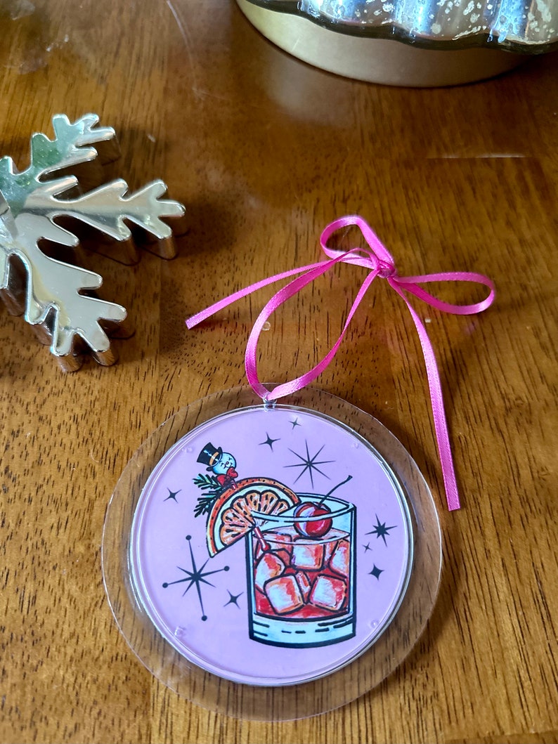 Pu&ograve; includere: Un addobbo natalizio rotondo rosa e trasparente con un'illustrazione di cocktail festivo. Il cocktail &egrave; una bevanda rossa con cubetti di ghiaccio, una ciliegia, una fetta d'arancia e un rametto di verde. Uno snowman con un cappello a cilindro &egrave; sullo sfondo.