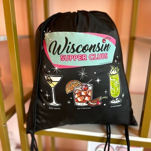 Puede incluir: Bolsa de cordón negra con un diseño retro que presenta el texto "Wisconsin Supper Clubs" e ilustraciones de tres cócteles: un Dirty Martini, un Old Fashioned y un Grasshopper.