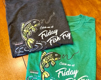 Può includere: Due magliette verde e grigia con un pesce che salta fuori dall'acqua e il testo "Catch me at Friday Fish Fry".