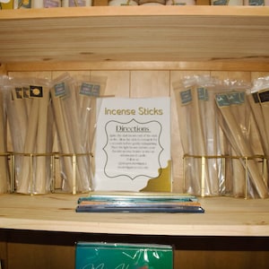 May include: A wooden shelf displays packaged incense sticks in clear plastic sleeves. Small chalkboard signs read "10ct Incense Sticks." A larger sign provides directions for use. The shelf is made of light-colored wood.