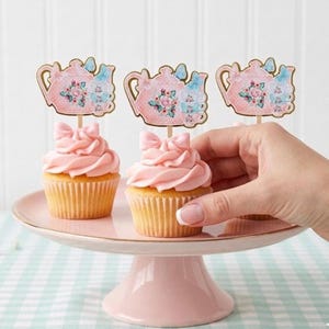 May include: Pink frosted cupcakes with bow-shaped frosting and decorative teapot toppers. The cupcakes are displayed on a pink cake stand. The toppers feature a pink teapot design with floral and butterfly accents.
