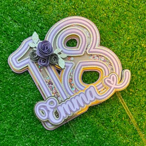 May include: Purple and gold layered paper cake topper with the number 18 and the name Emma. The number 18 has a glittery texture and the name Emma is in a cursive font.