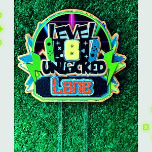 May include: A green, blue, yellow, and orange cake topper with the text "Level 8 Unlocked" and the name "Lane".